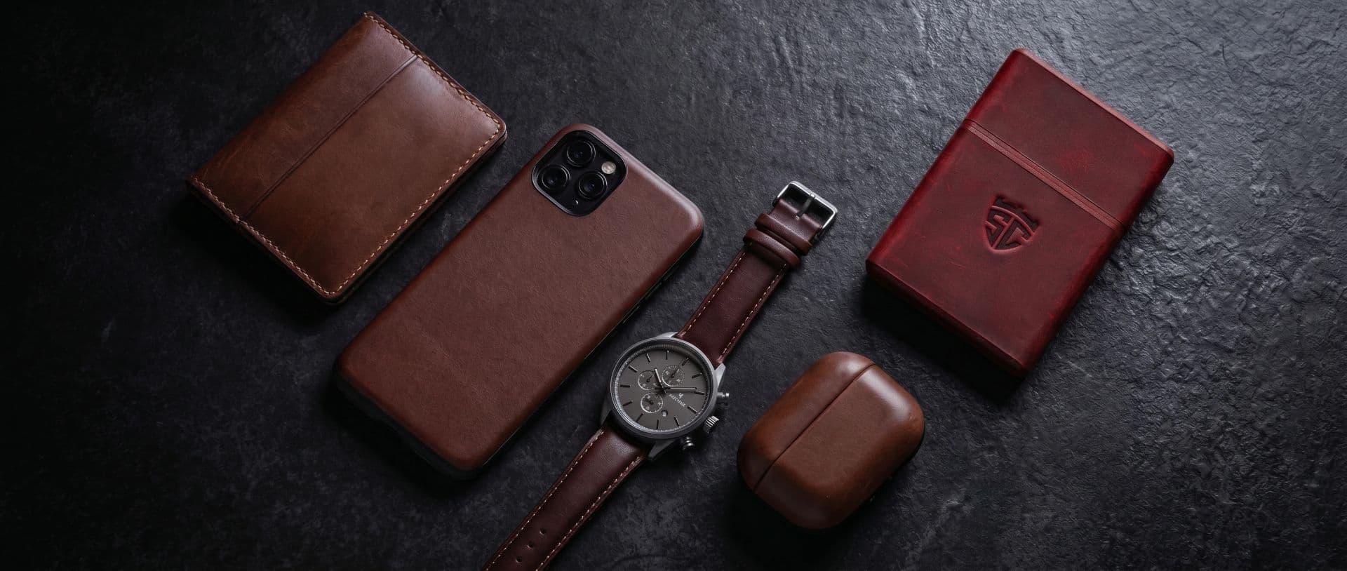 Leather wallet, phone case, watch strap and a red cigarette case on dark stone